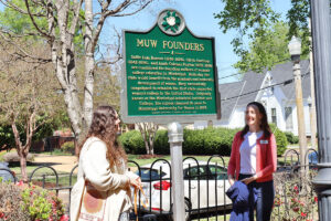 Two women stand on either side of a tall green historical marker labeled MUW Founders