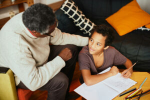 An older man leans over and helps a young kid with their homework