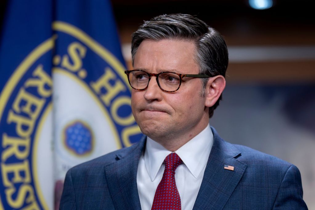 A man in blue suit and red tie grimaces before a flag bearing the seal of the US House of Representatives