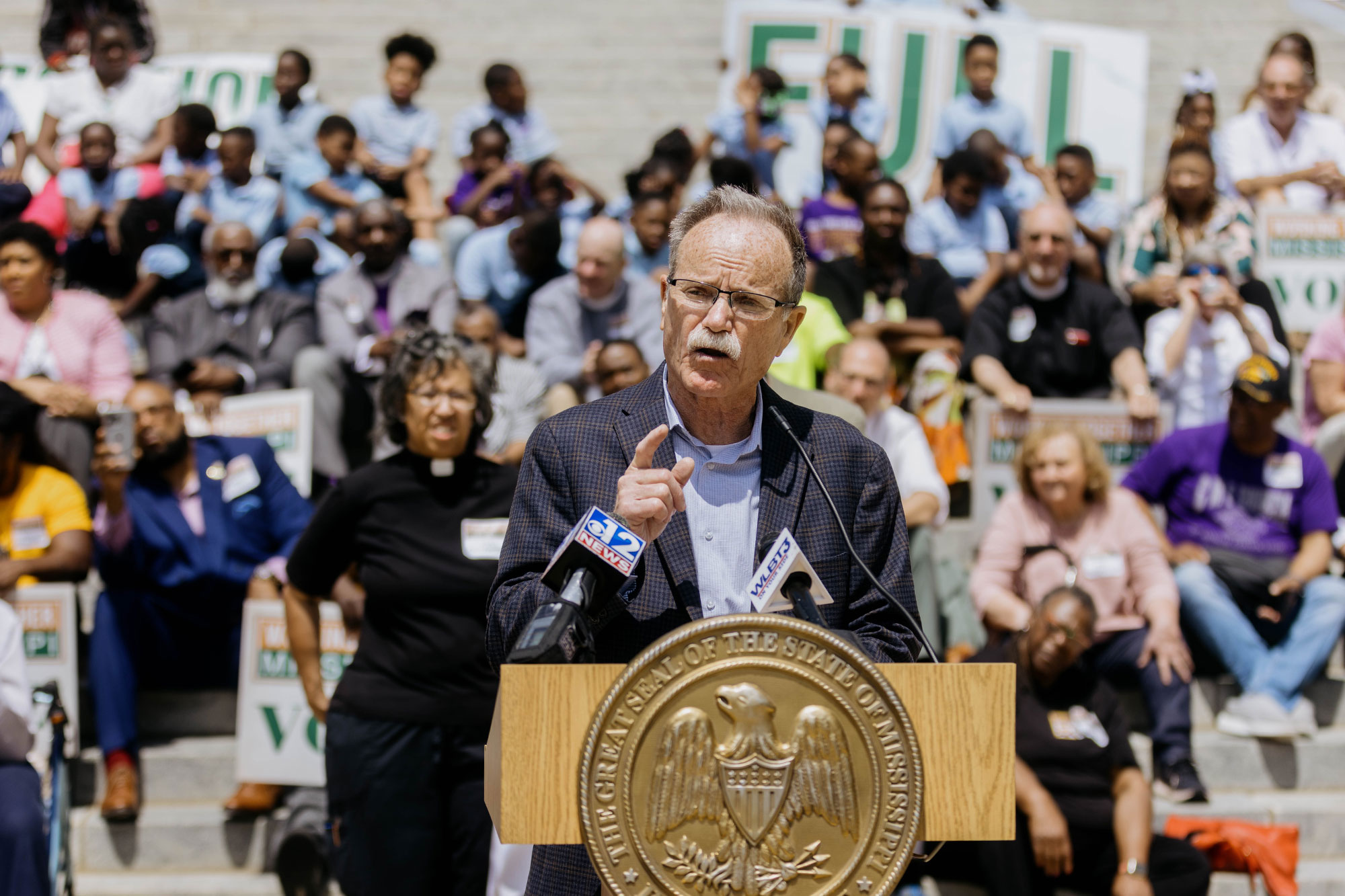 A white man speaks before a crowd at a podium bearing a golden Mississippi state seal