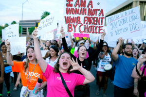 Protestors march down the road holding signs. The most center sign reads "I am a woman not a womb. Parent by choice."