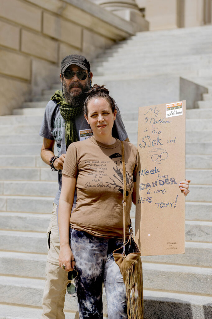 A man and a woman pose by a tall set of stone stairs. The woman is holding a sign that says "My mother is too sick and weak with cancer to come here today!"