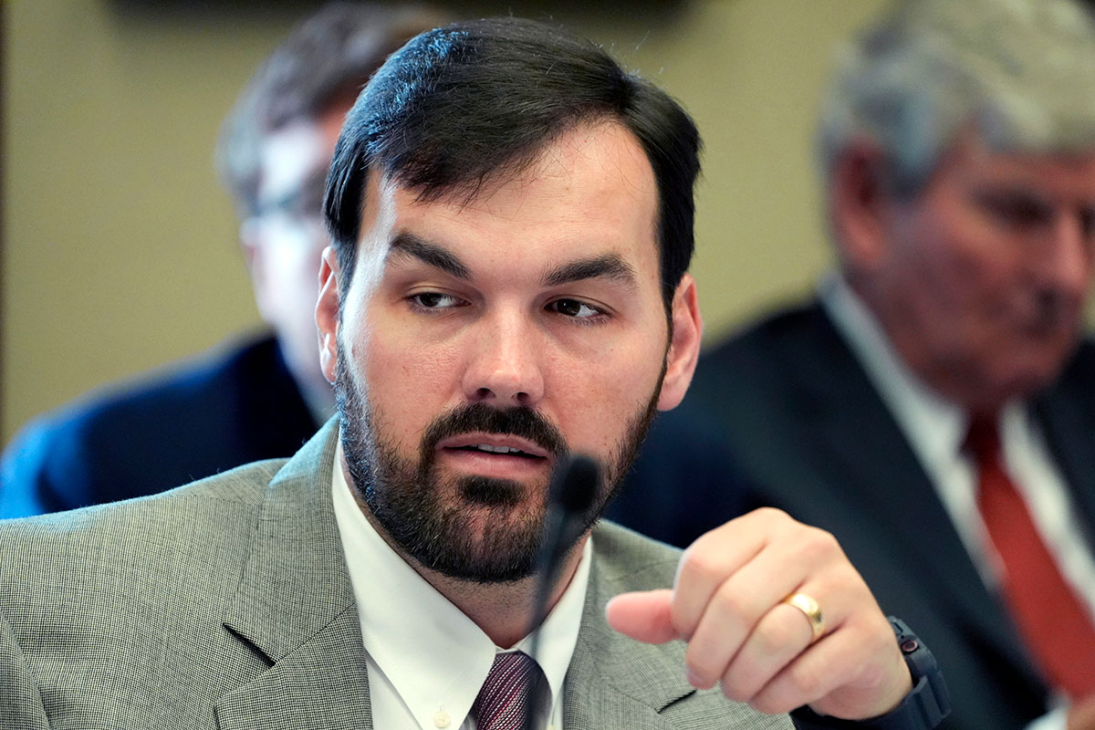 A man seated in a committee meeting with others. He's wearing a grey suit and striped tie, and his upraised hand shows a gold wedding band.
