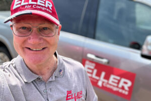 A man wearing a red hat that reads Ron Eller for Congress, plus a grey shirt with Ron Eller for Congress embroidered in red thread, poses in front of a grey vehicle with a red Ron Eller for Congress magnet on the door