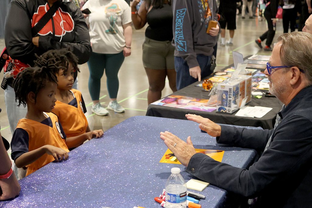 Two children dressed in bright orange outfits from Dragon Ball stand on the other side of the table from the voice actor for Goku