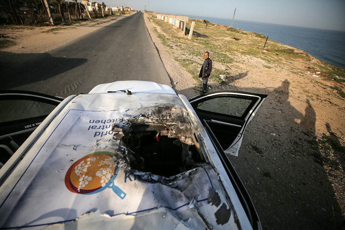 A man looks towards a white vehicle with a giant bomb hole in the roof