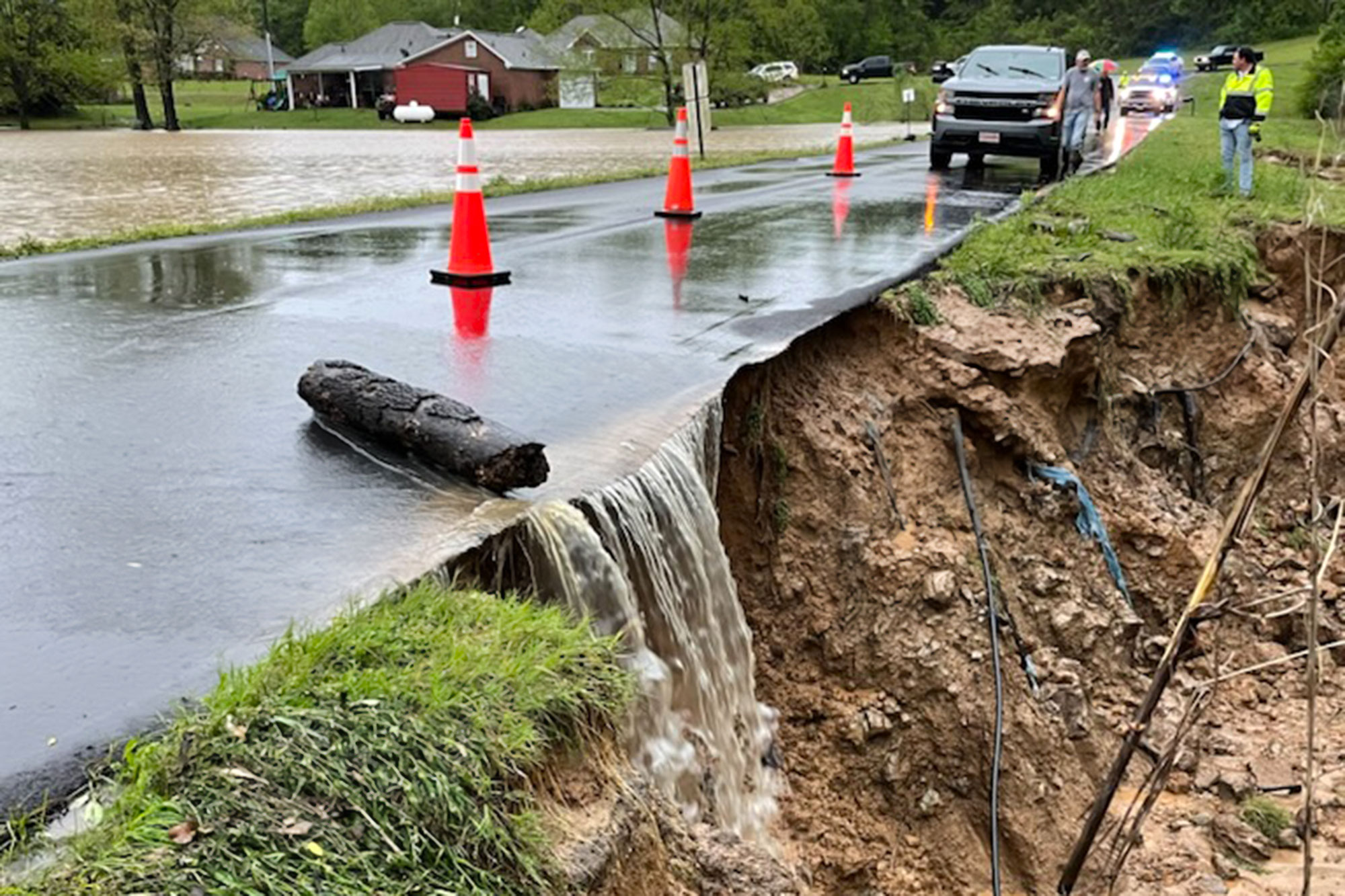 A flooded road blocked with orange cones. Bare dirt is exposed on the side and water is spilling over