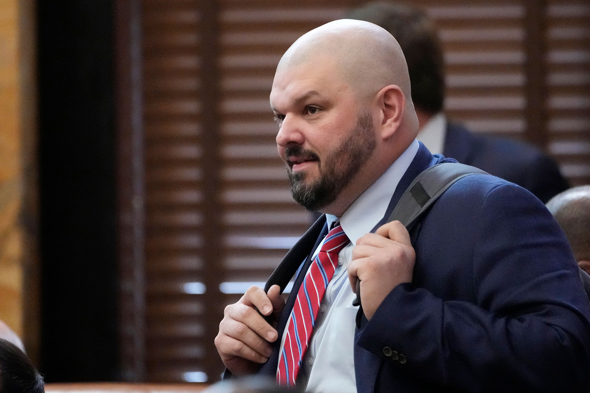 A bearded, bald man wearing suit and tie holds backpack straps at his chest and faces the left