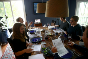 5 people sit at a dining room table, the end sixth seat taken by a laptop displaying a zoom screen with several more people