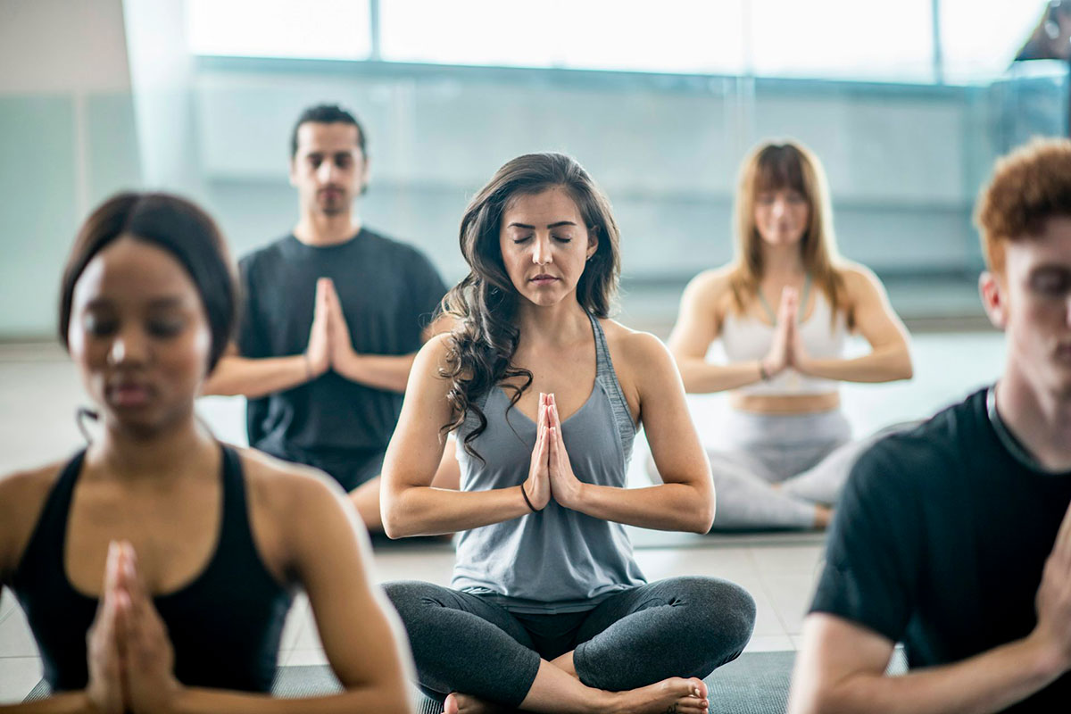 A yoga class sits cross-legged on matts and hold their hands together as though in prayer