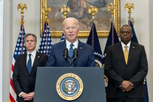 President Biden speaks from a podium branded with the President of the United States seal on front. Two men flank behind him