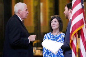 A man in a black suit, left, speaks to a woman in a blue and white dress and another man in grey suit slightly obstructed by an American flag in the foreground