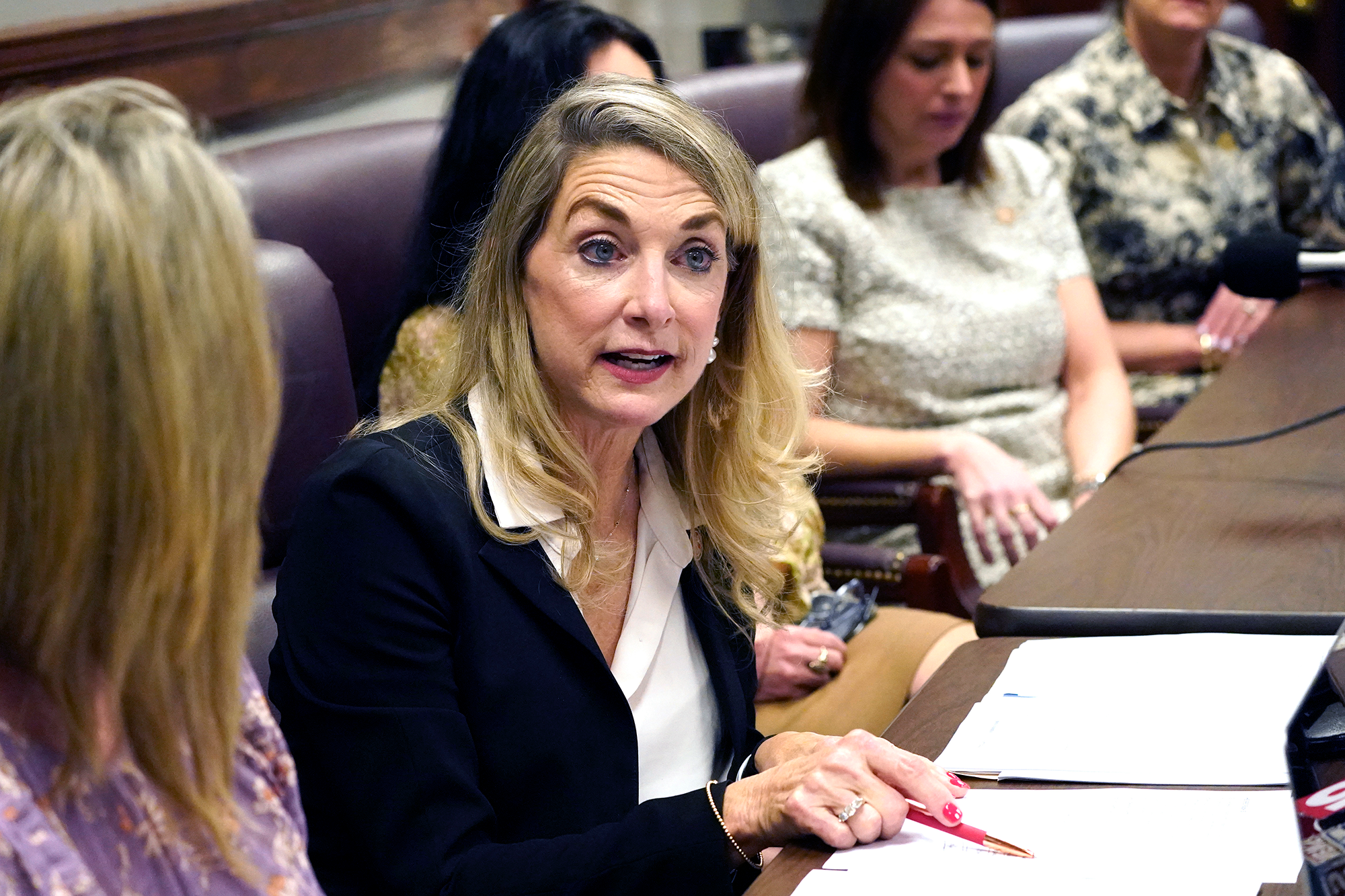 A blonde woman in a suit speaks while gesturing to a print out with a pen in hand
