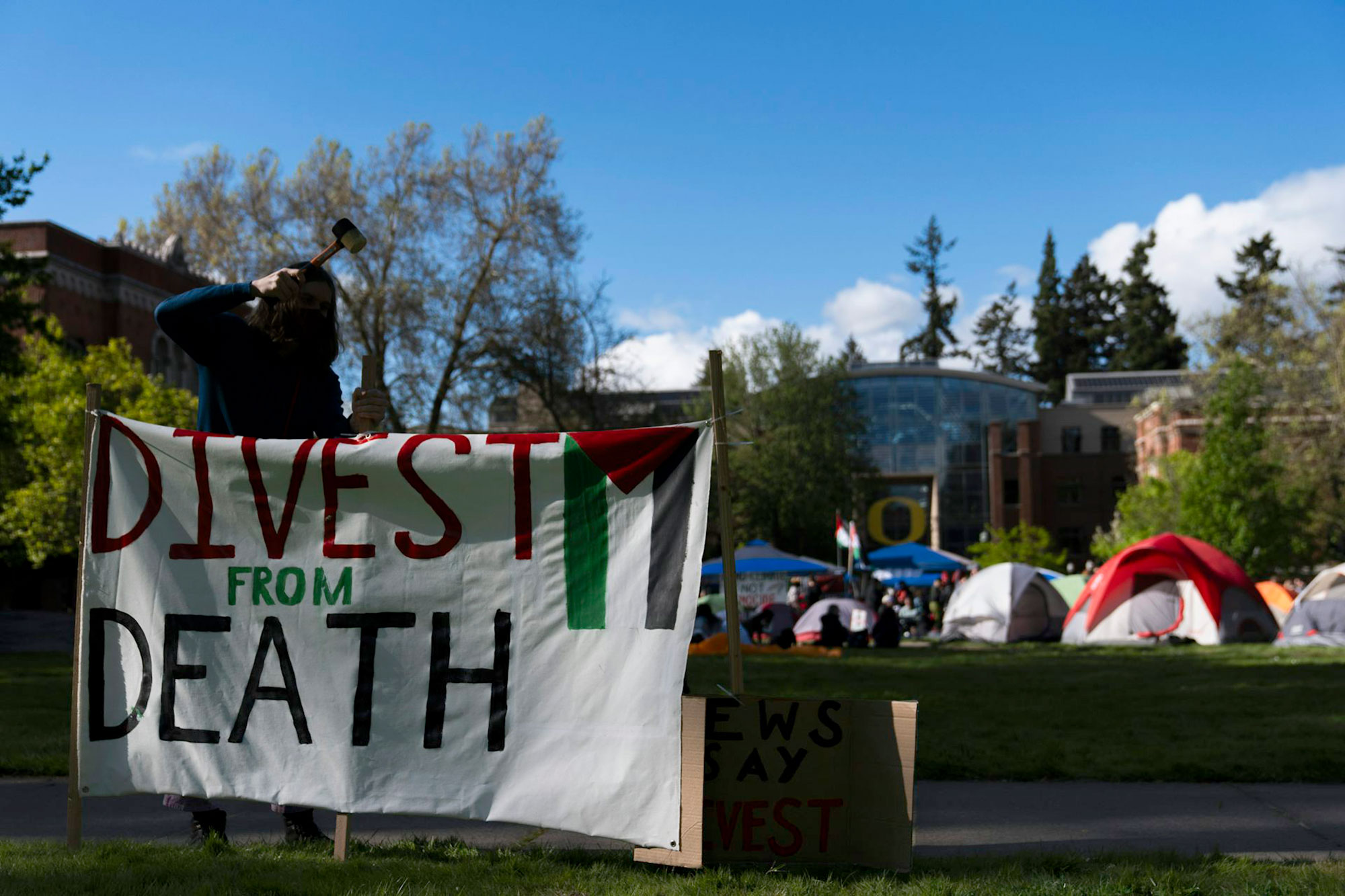 A person is hanging a banner that reads "Divest from Death". Several tends are seen in the campus background