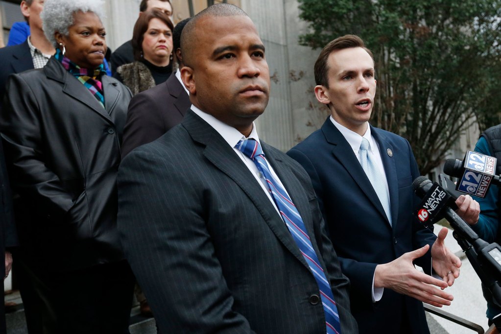 Two mean dressed in suits speak at a podium outside of a building. Others stand further behind them.
