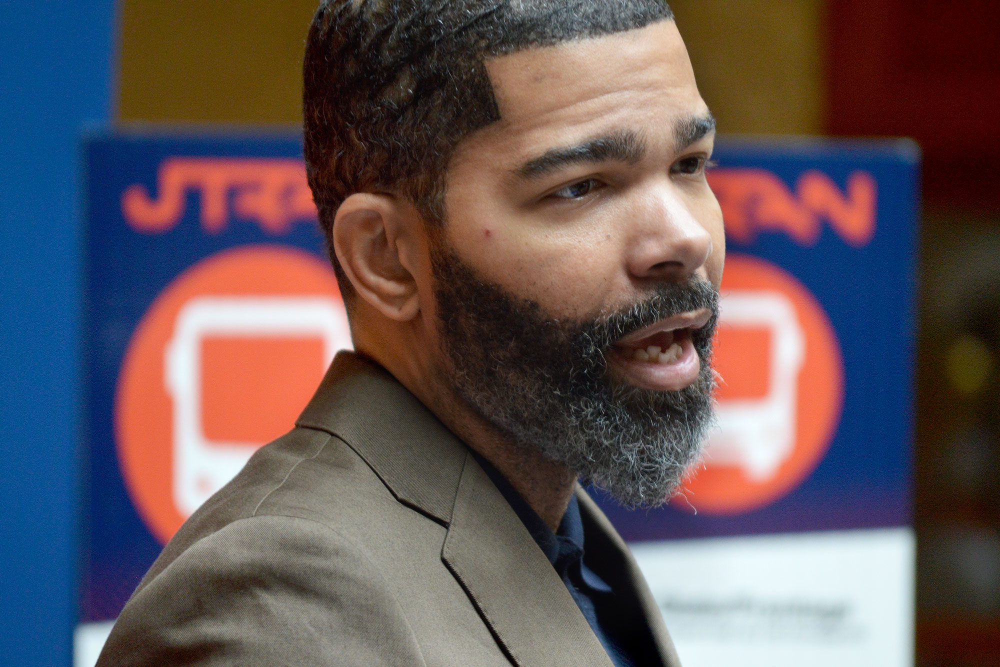 A bearded man in a tan suit speaks. A blue and orange sign that reads JTRAN is visible behind him