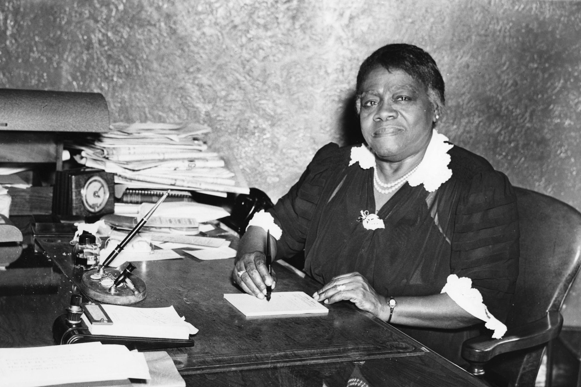 A woman sits at a busy writing desk with pen in hand