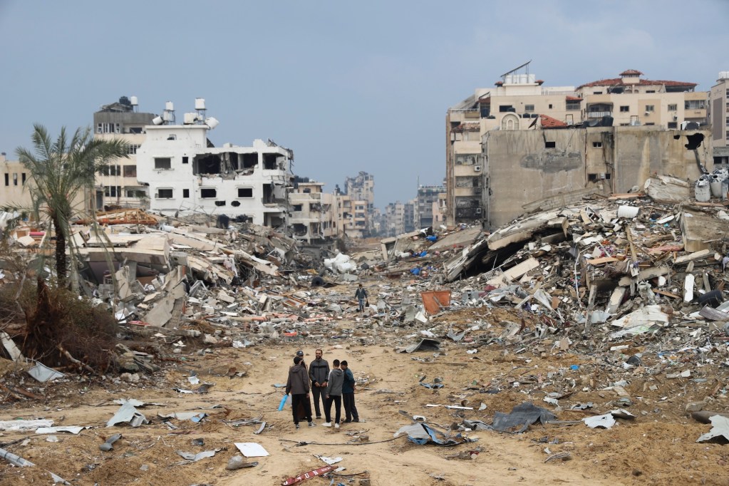 a photo shows four people standing in the ruins of a bombed out Gaza neighborhood