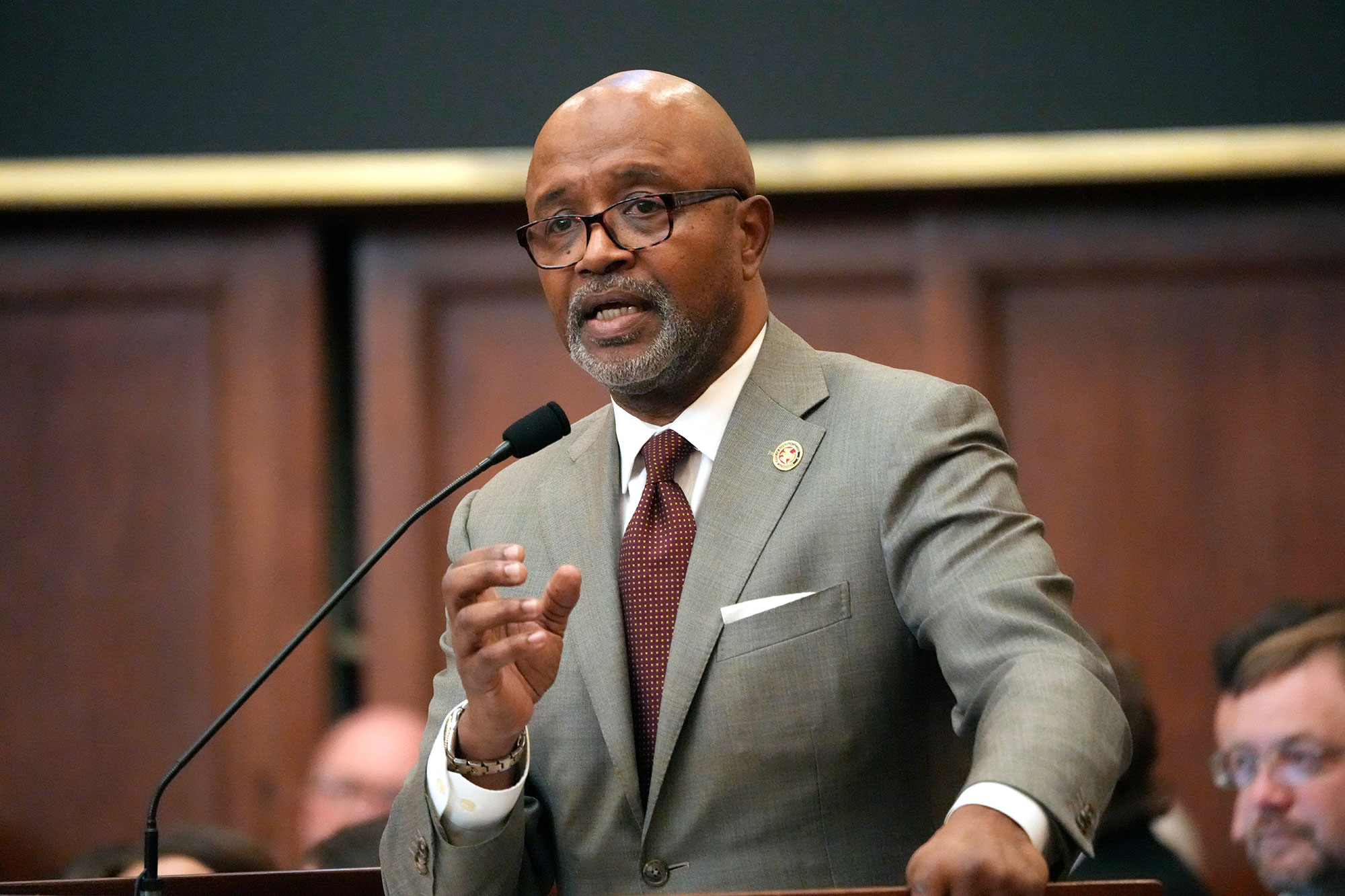 A man in a glasses, a grey suit and brown tie speaks at a podium while gesturing with one hand