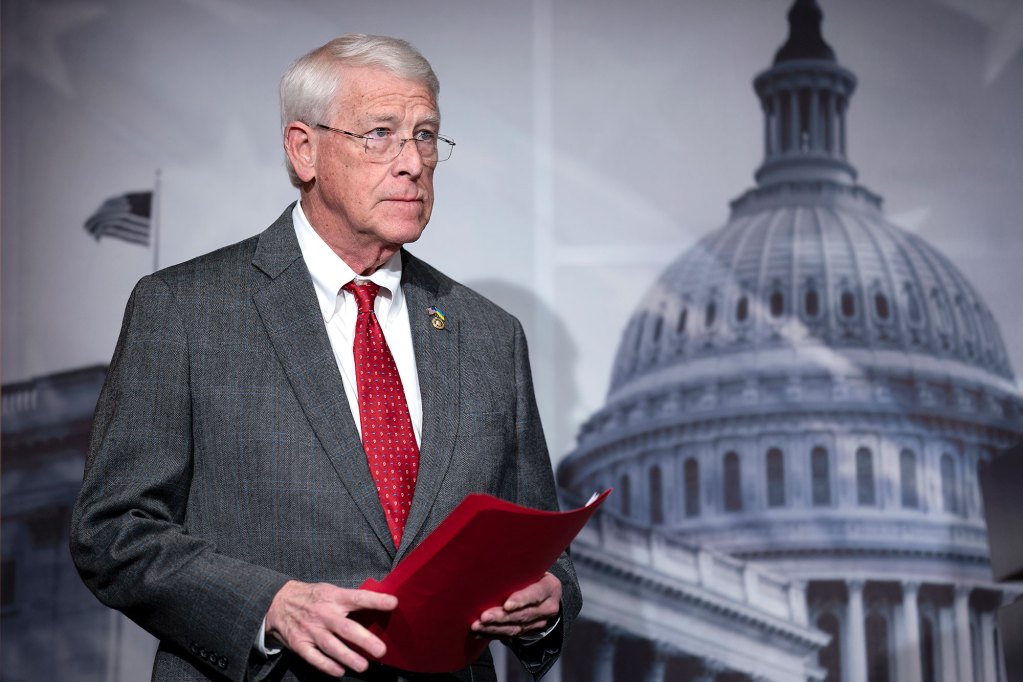 A man in grey suit and red tie stands in front of a print of the Miss. Capital building