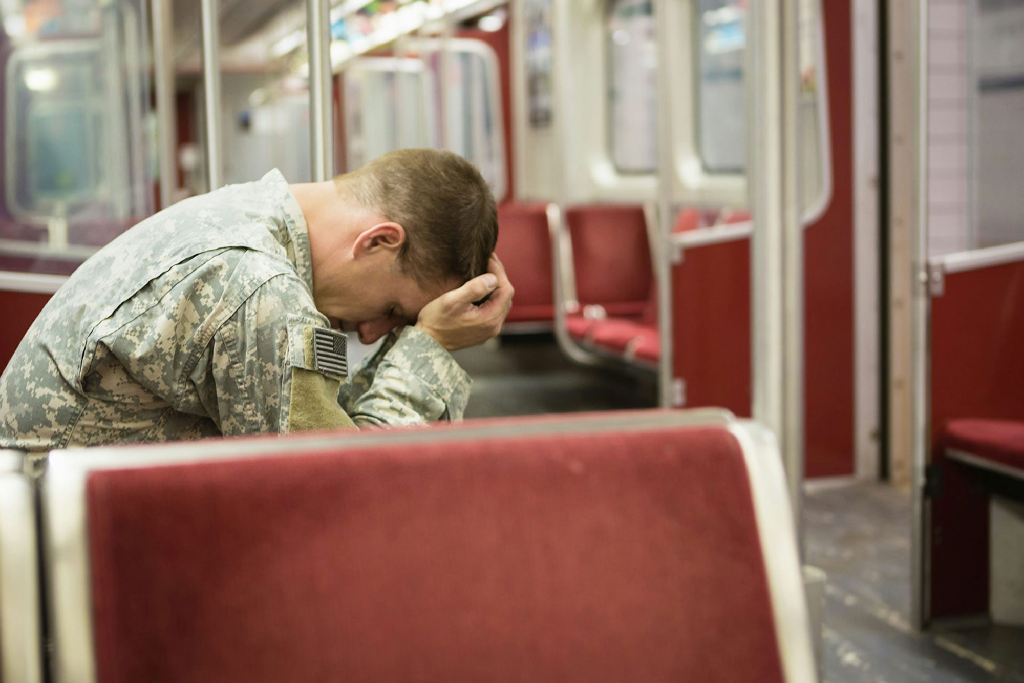 A person in military uniform camouflage sitting on a train rests their head in their hands