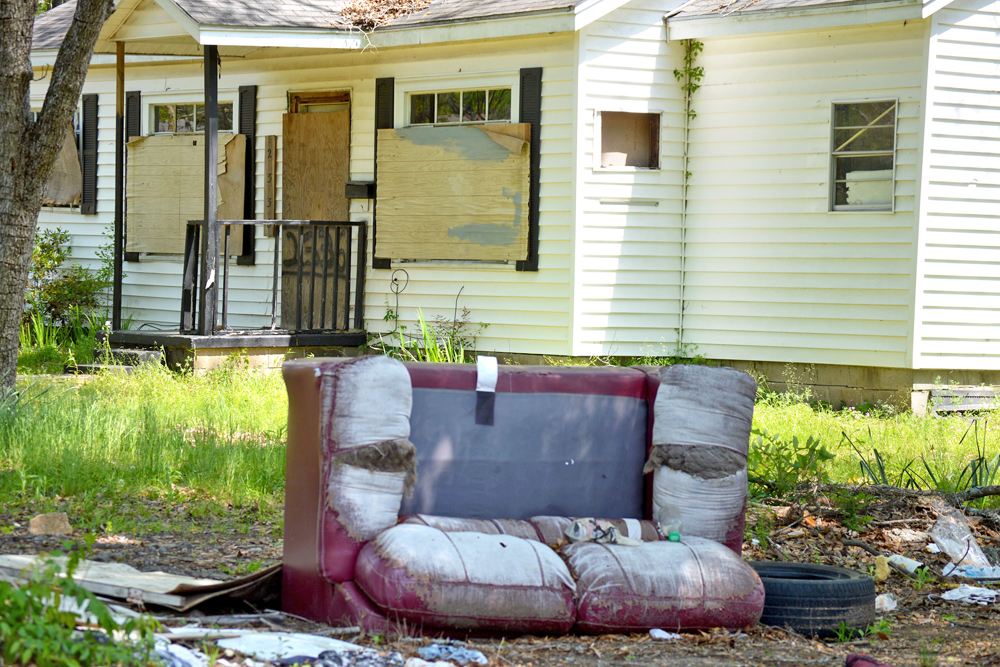 A boarded up white house with debris in the yard including a large worn out red couch and tires