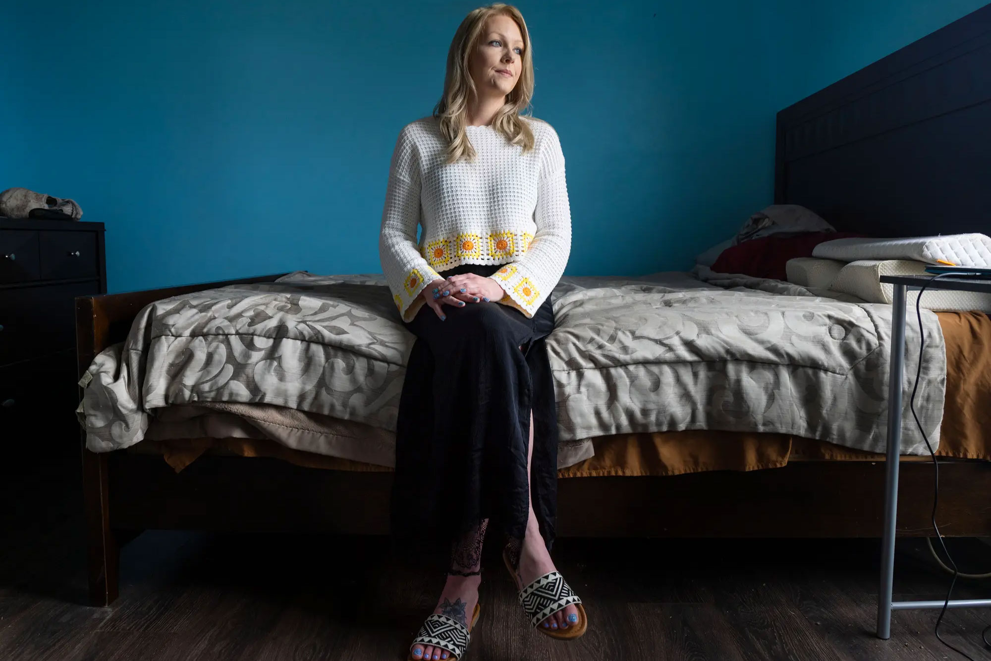 A blonde woman sits on a bed in a room with blue walls