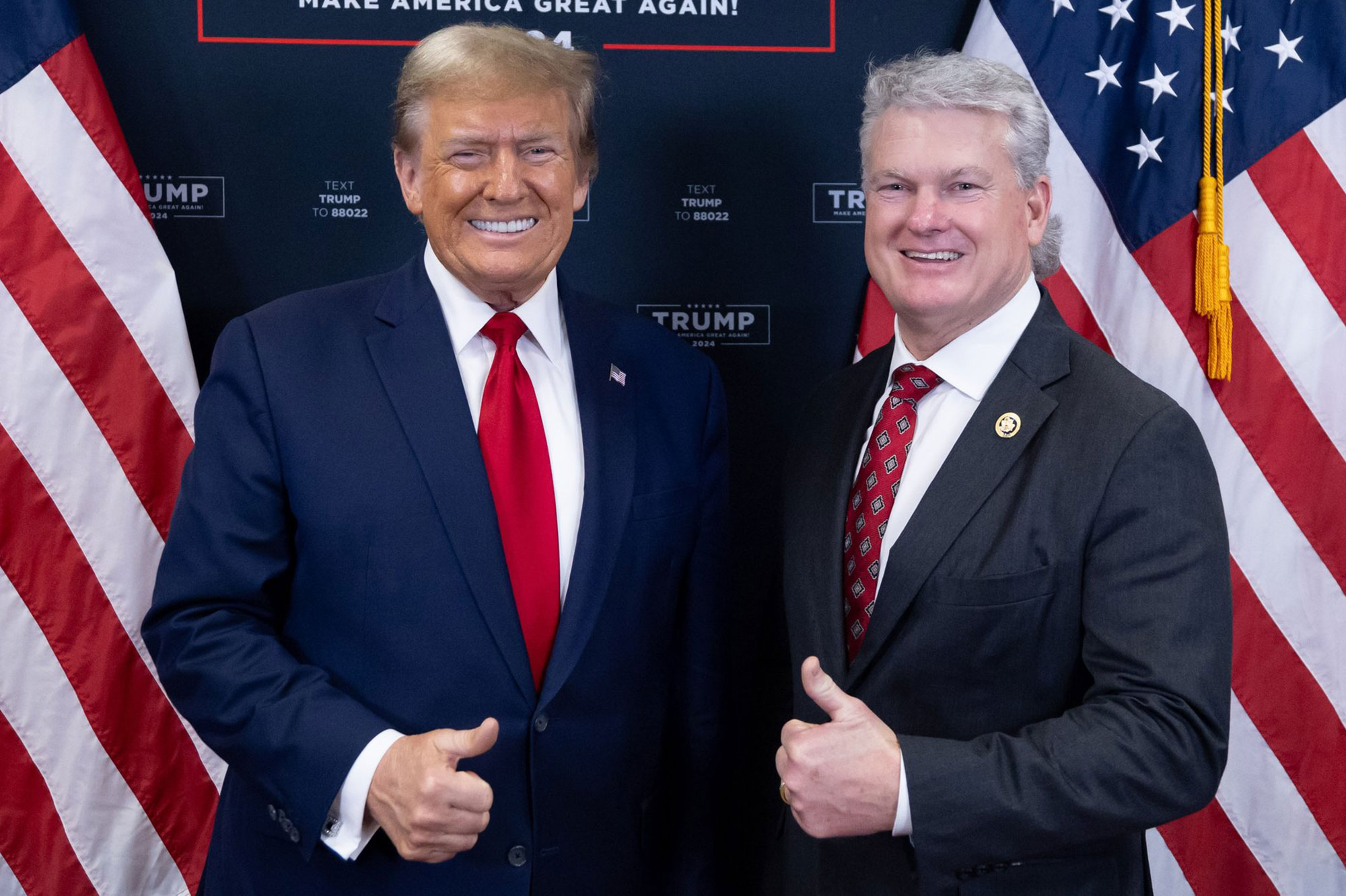 Former President Donald Trump and Representative Mike Collins do a posed thumbs up before a Trump wall and US Flags