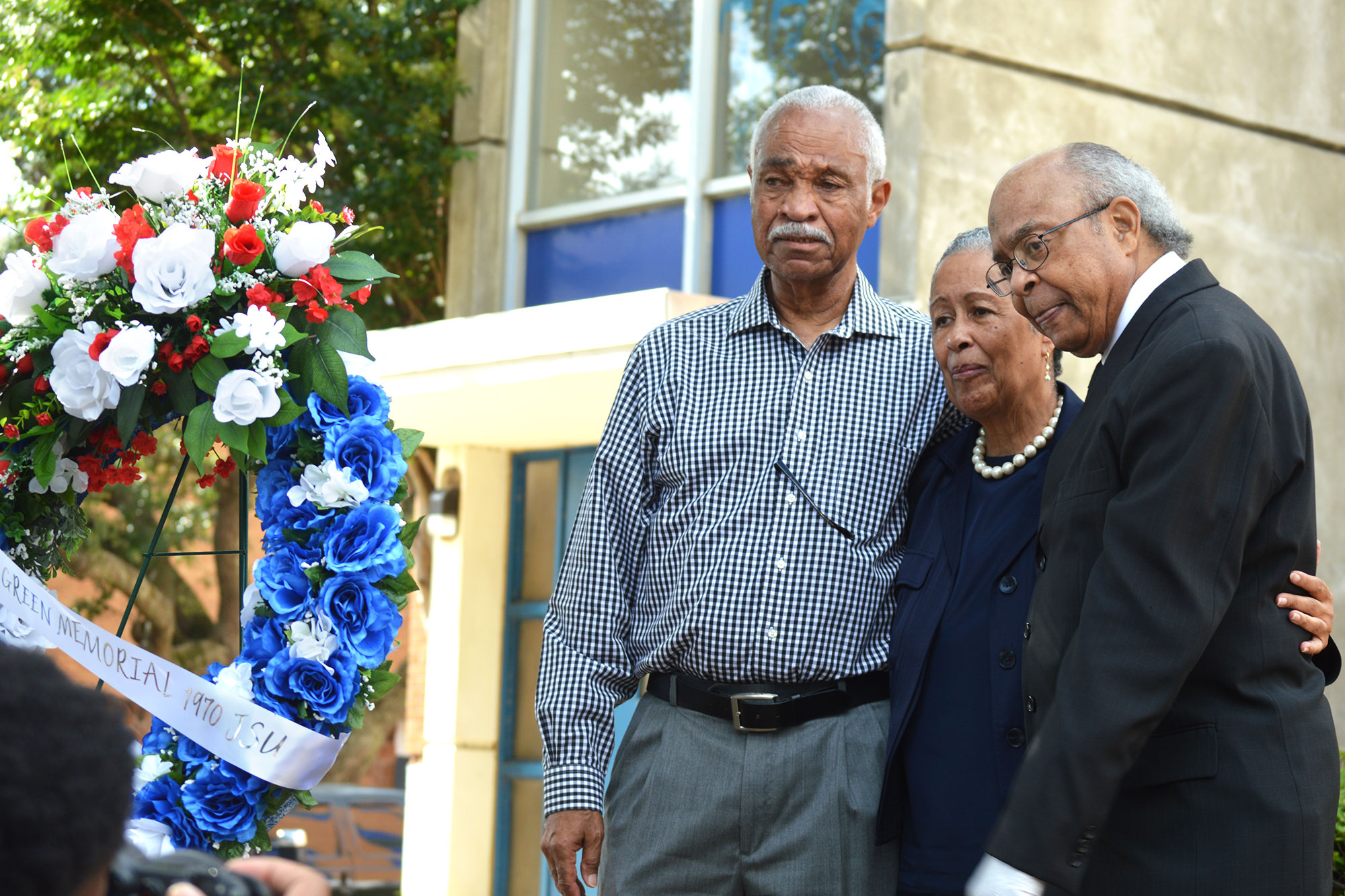 Three JSU alumni hug close and look on at a commemorative wreath made of blue and white roses