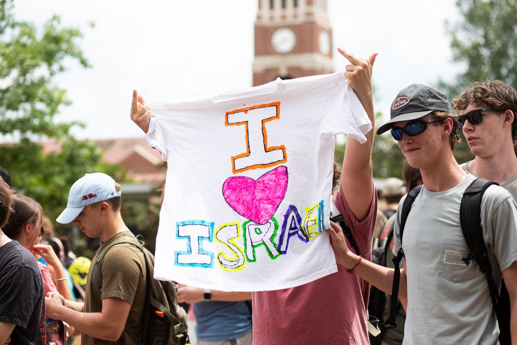 a white male student holds up an "I Heart Israel" shirt while flipping the bird with both hands