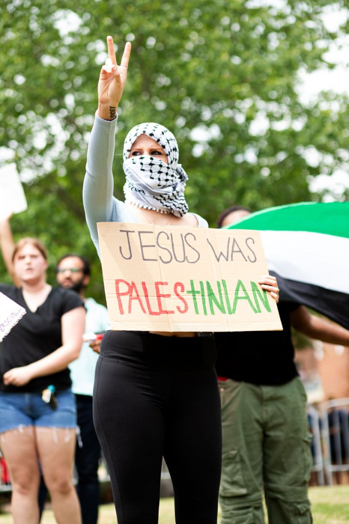 a woman holds a Jesus was a palestinian sign while holding up a peace sign with her fingers