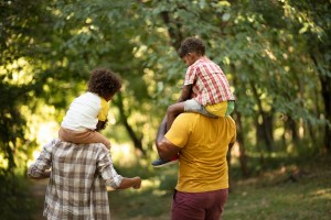 Two adults carry two small children on their shoulder and walk through a sunlit wooded area