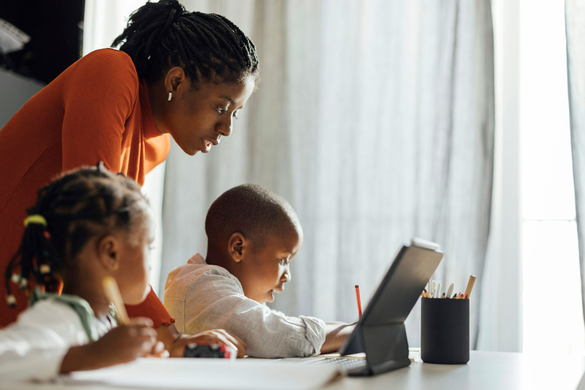 A woman in an orange shirt leans over two younger children working on class work.
