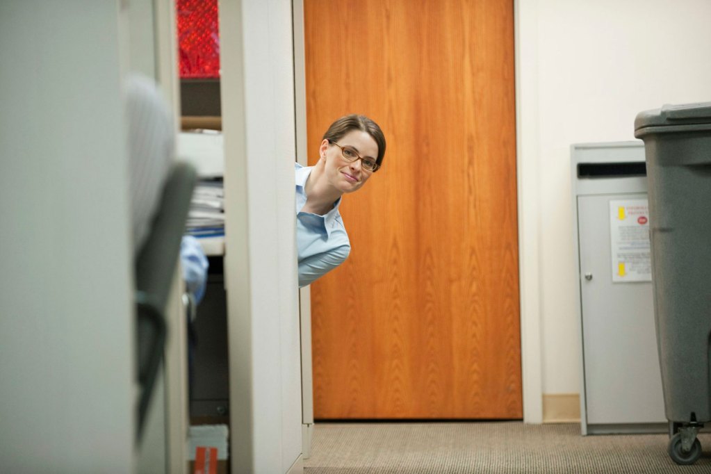 A brunette woman in glasses peeks around a wall of an office, looking at the photographer.