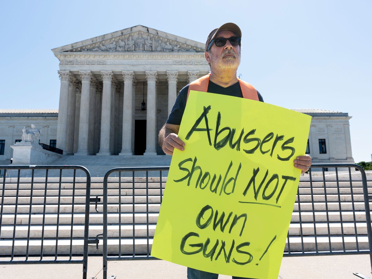 A man stands outside of the U.S. Supreme Court holding a bright yellow sign that says "Abusers should NOT own guns"