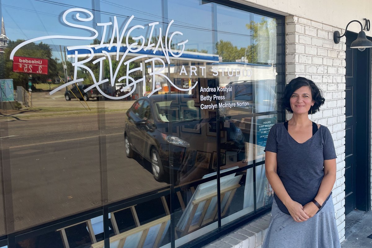 A woman posed outside of a white brick storefront. The window is labeled "Singing River Art Studio"