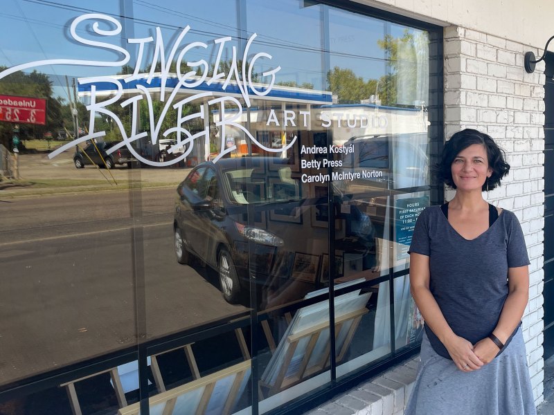 A woman posed outside of a white brick storefront. The window is labeled "Singing River Art Studio"