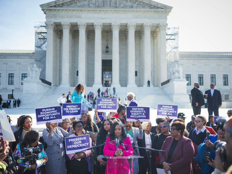A woman in bright pink suit speaks before a crowd standing in front of the U.S. Supreme Court. The people hold signs that say Pursue Economic Opportunity for All