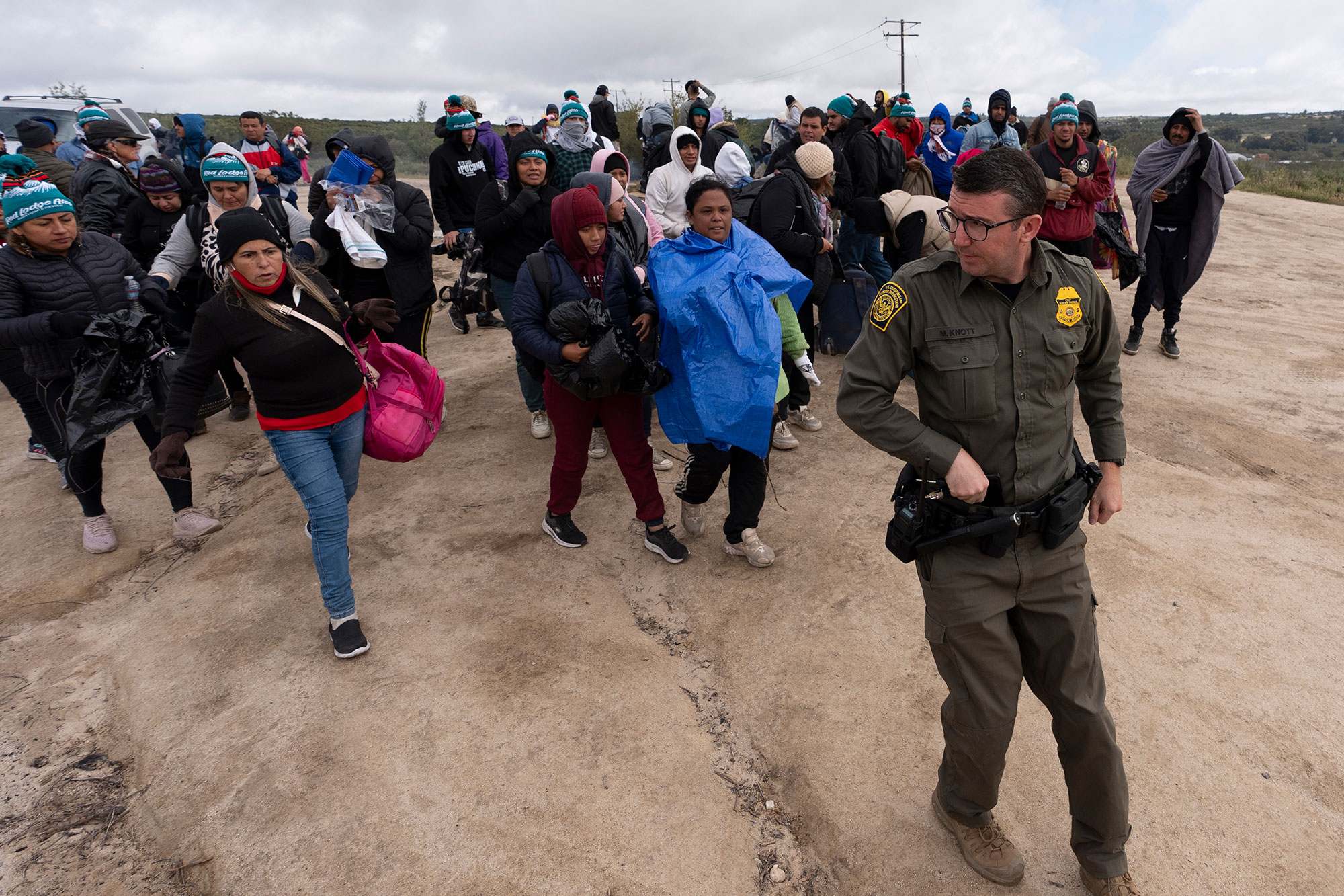 eople seeking asylum, including a group from Peru, walk behind a Border Patrol agent