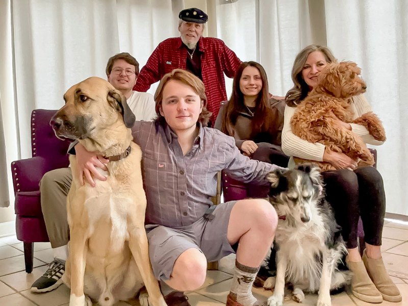 A family of three men and two women sit on a large purple couch while holding three dogs between them