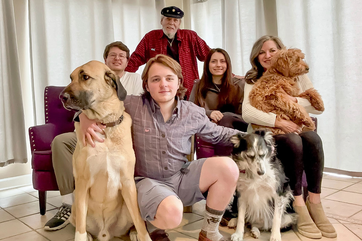 A family of three men and two women sit on a large purple couch while holding three dogs between them