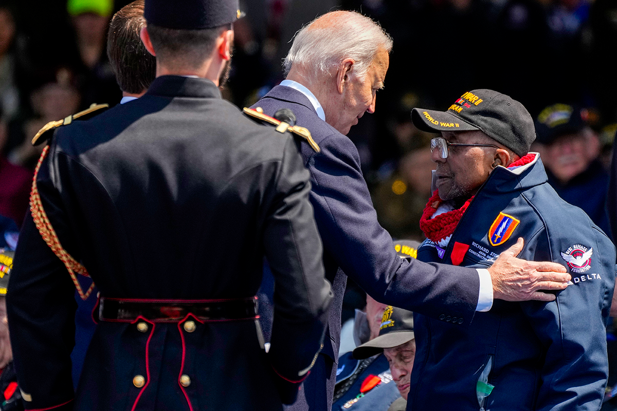 A man sets his arm on the shoulder of an older man in WWII veteran uniform, as a man in dress uniform looks on
