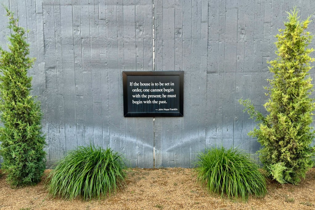 A small framed quote hung on a fence that reads "If the house is to be set in order, one cannot begin with the present; he must begin with the past." - John Hope Franklin