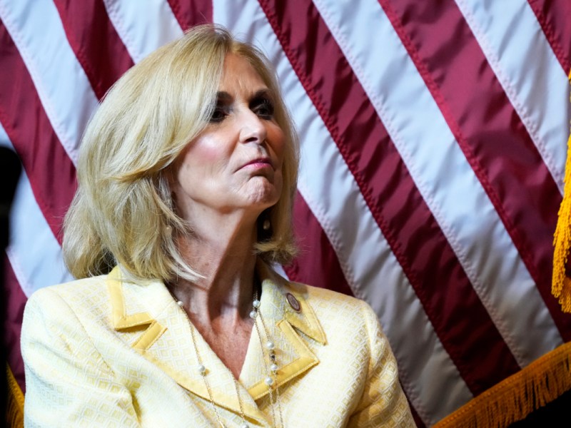 A woman in a yellow suit stands before the US Flag
