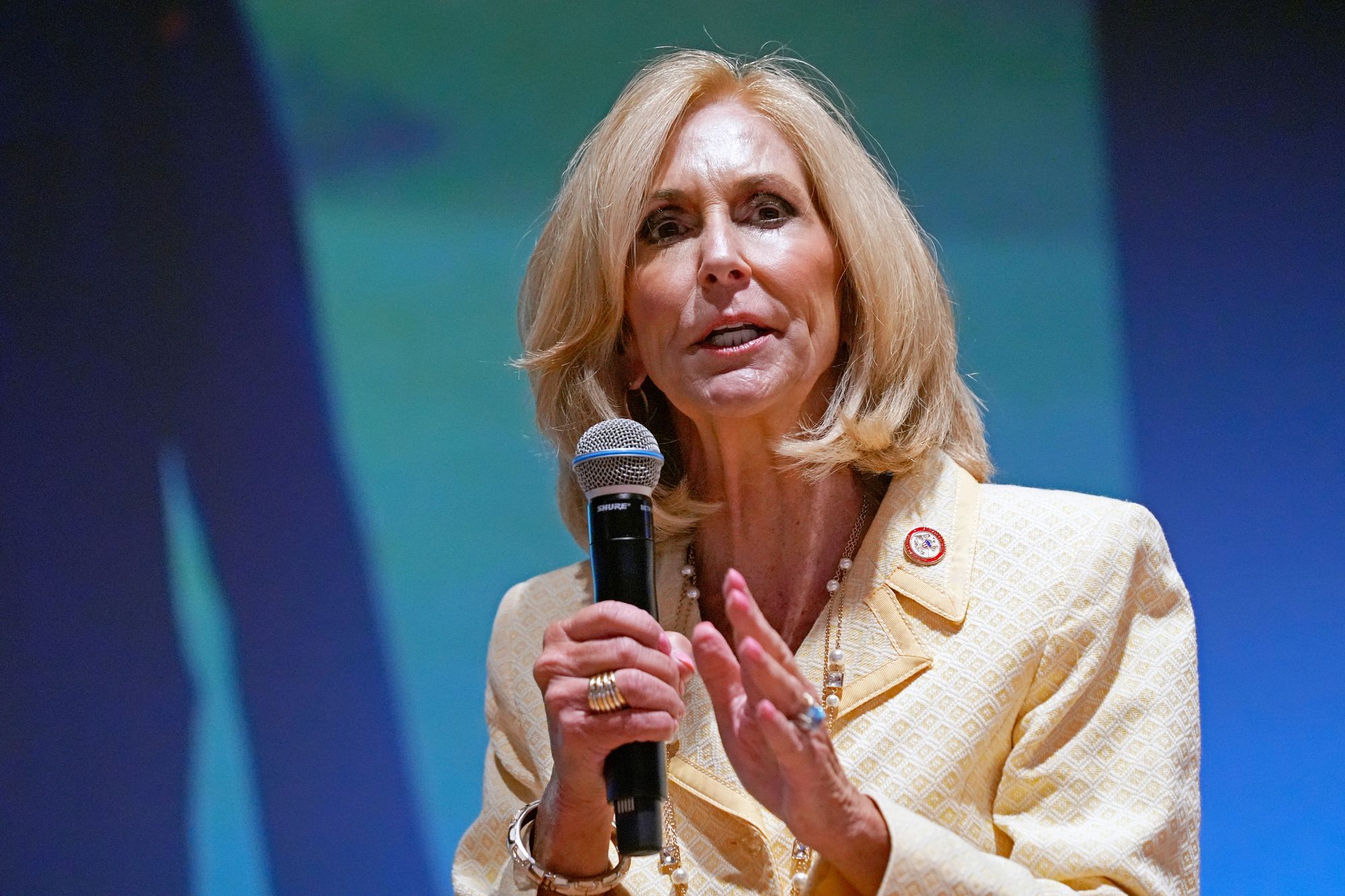 A blonde woman in a yellow outfit speaks on a stage that's backlit with a blue screen