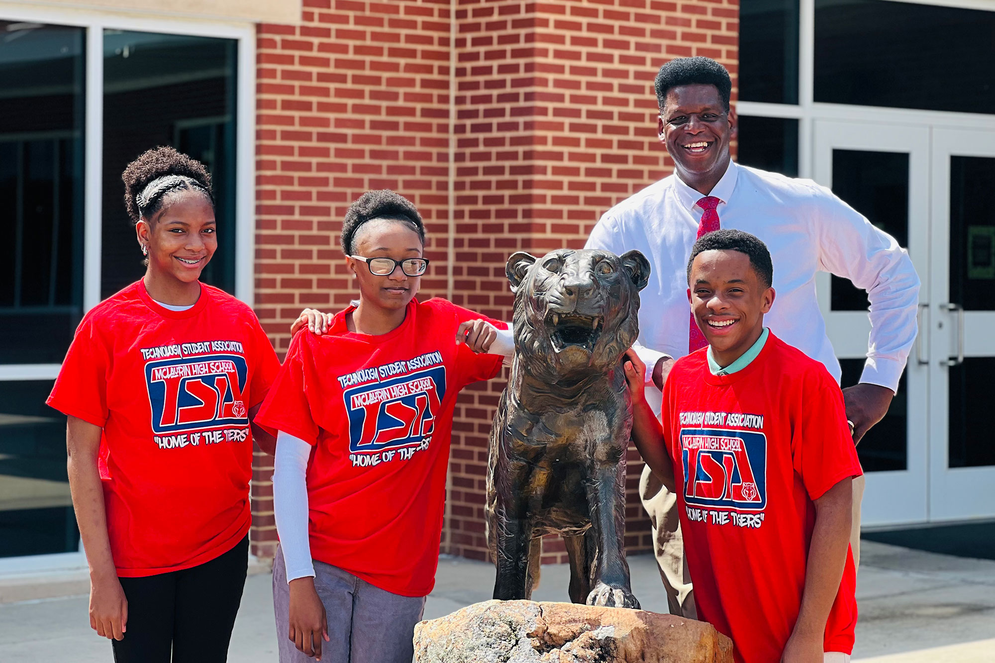 Three students in matching red tshirts and a teacher behind them post by the statue of a tiger