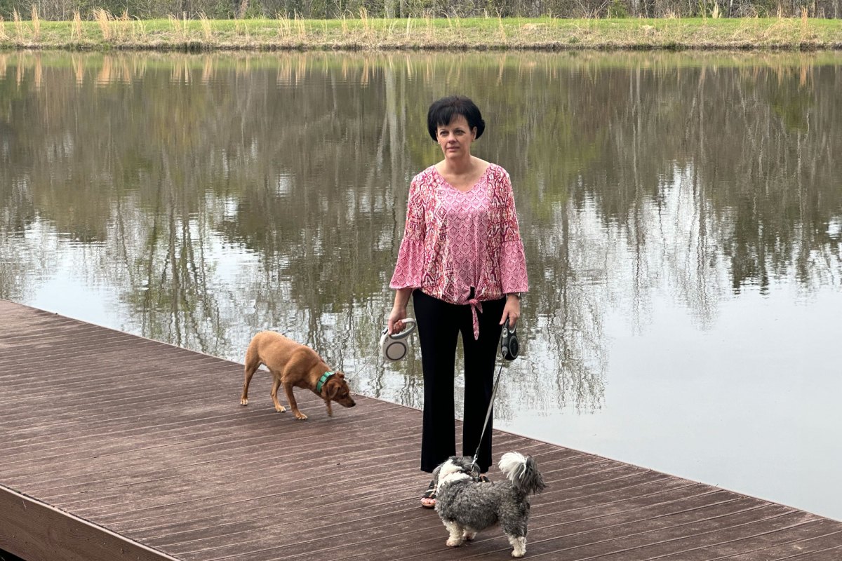 A woman stands on a lake pier while holding two small dogs by their leashes