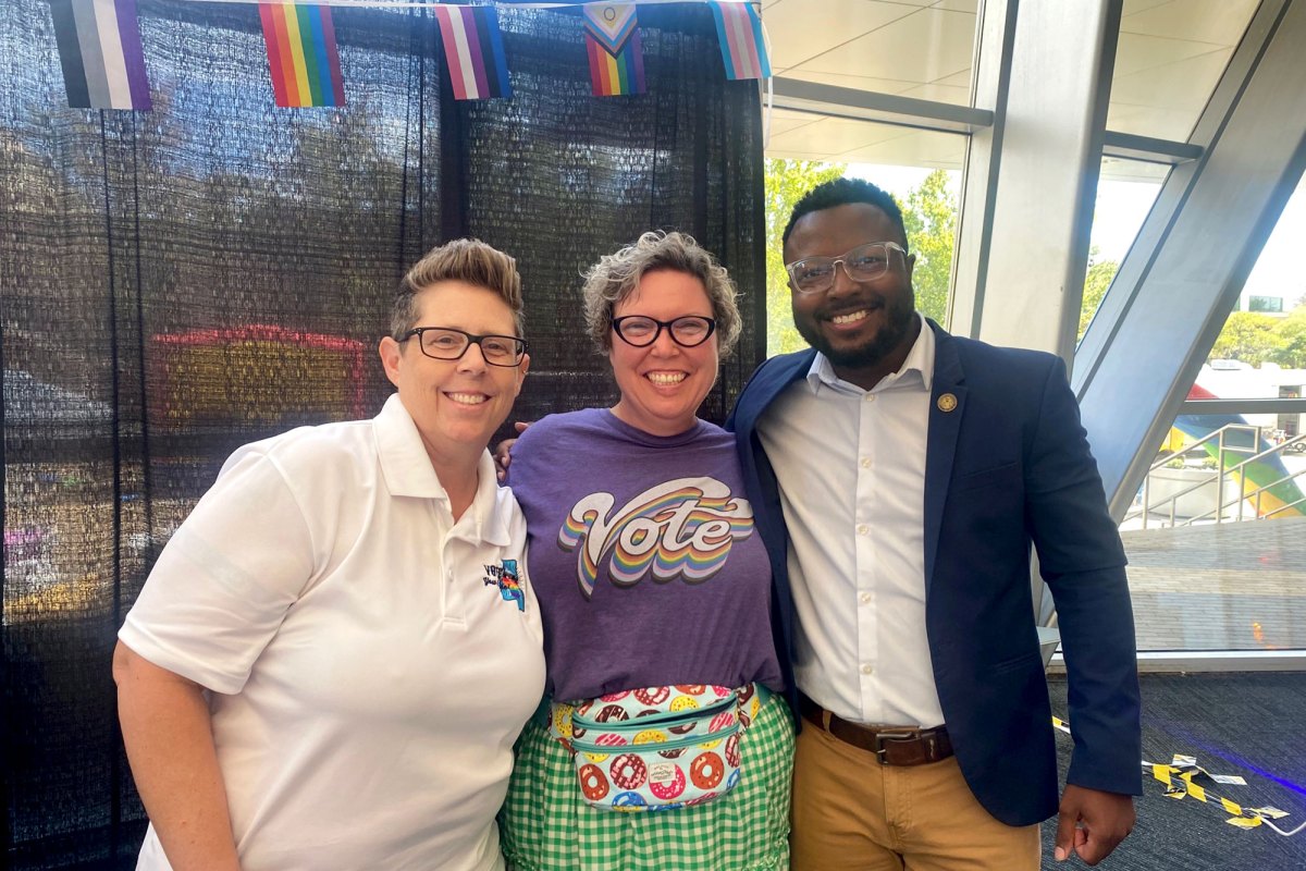 Three people pose together, the center person wearing a shirt that says "VOTE" in rainbow colors