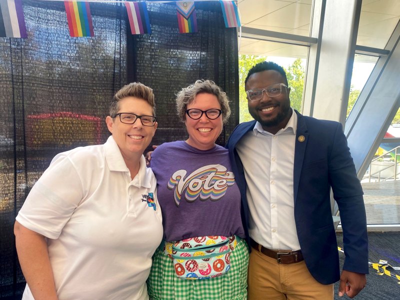 Three people pose together, the center person wearing a shirt that says "VOTE" in rainbow colors