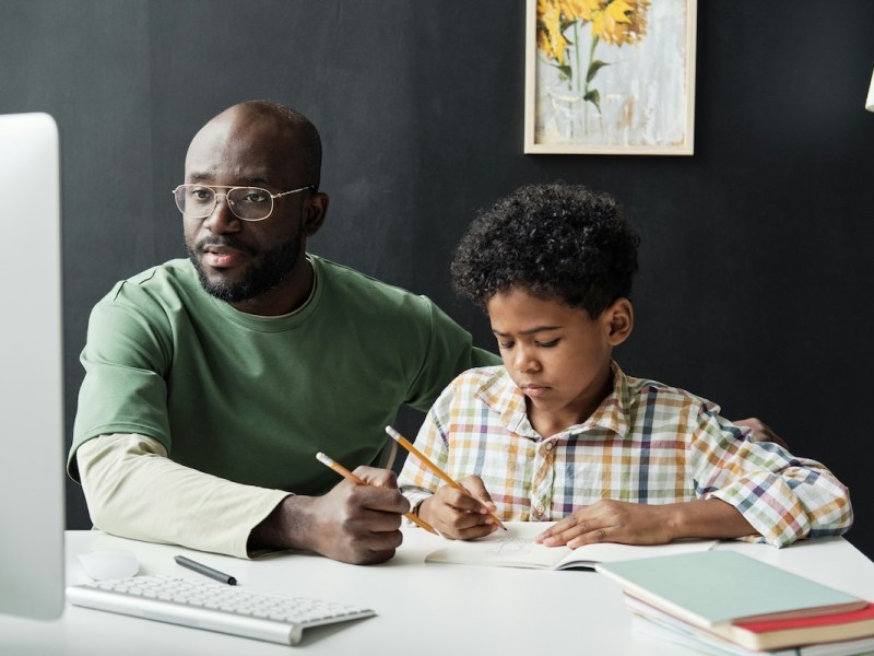A man looks on at a computer screen with pencil in hand helping a child with homework